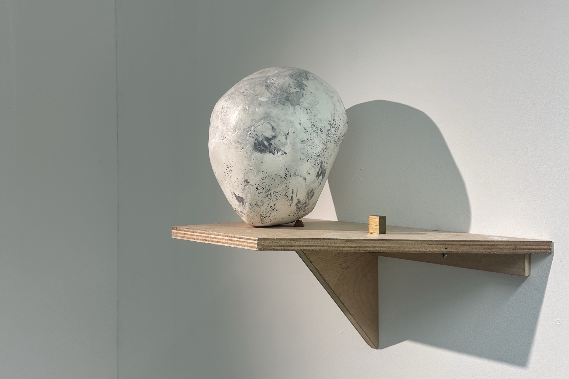 A black and white, marble looking head-like object, balanced on a small brass cube, standing on a wooden shelf. White wall in the background.
