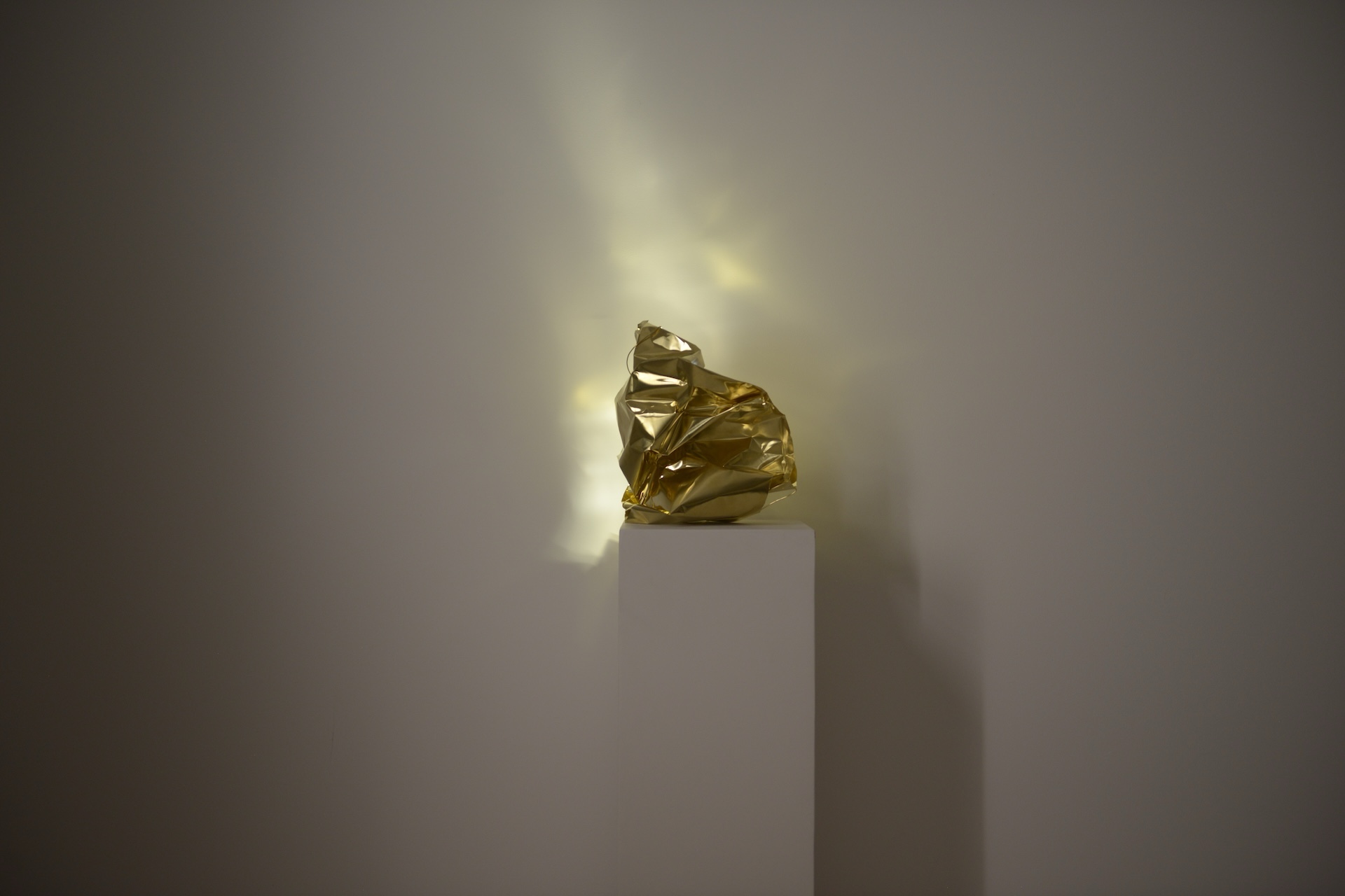A crimpled, polished brass object on top of a white plinth. Different shades of white light emerge from the inside and project an abstract shape on the wall behind it.