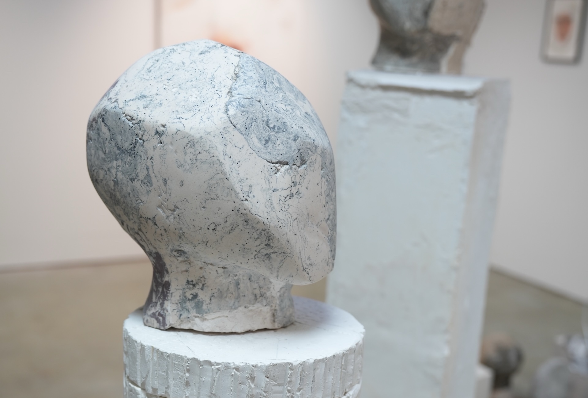 A stone-looking, polygonal head-like object placed on a white socle. Blurred plinth and other sculptures in the background.