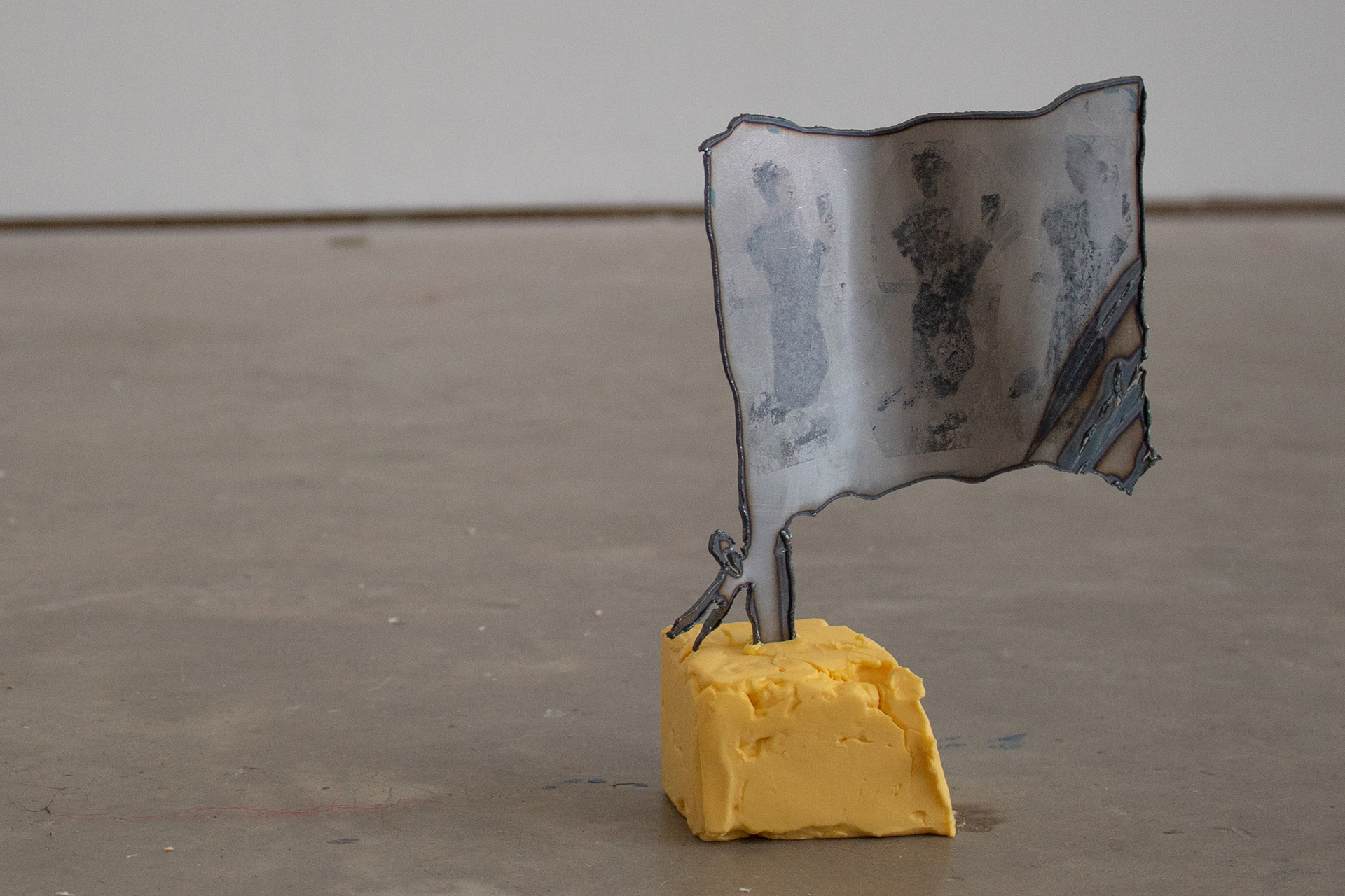 A small plasma cut steel flag with three figures photo-transferred on it is held upright by half a block of butter. The sculpture stands on a concrete floor.