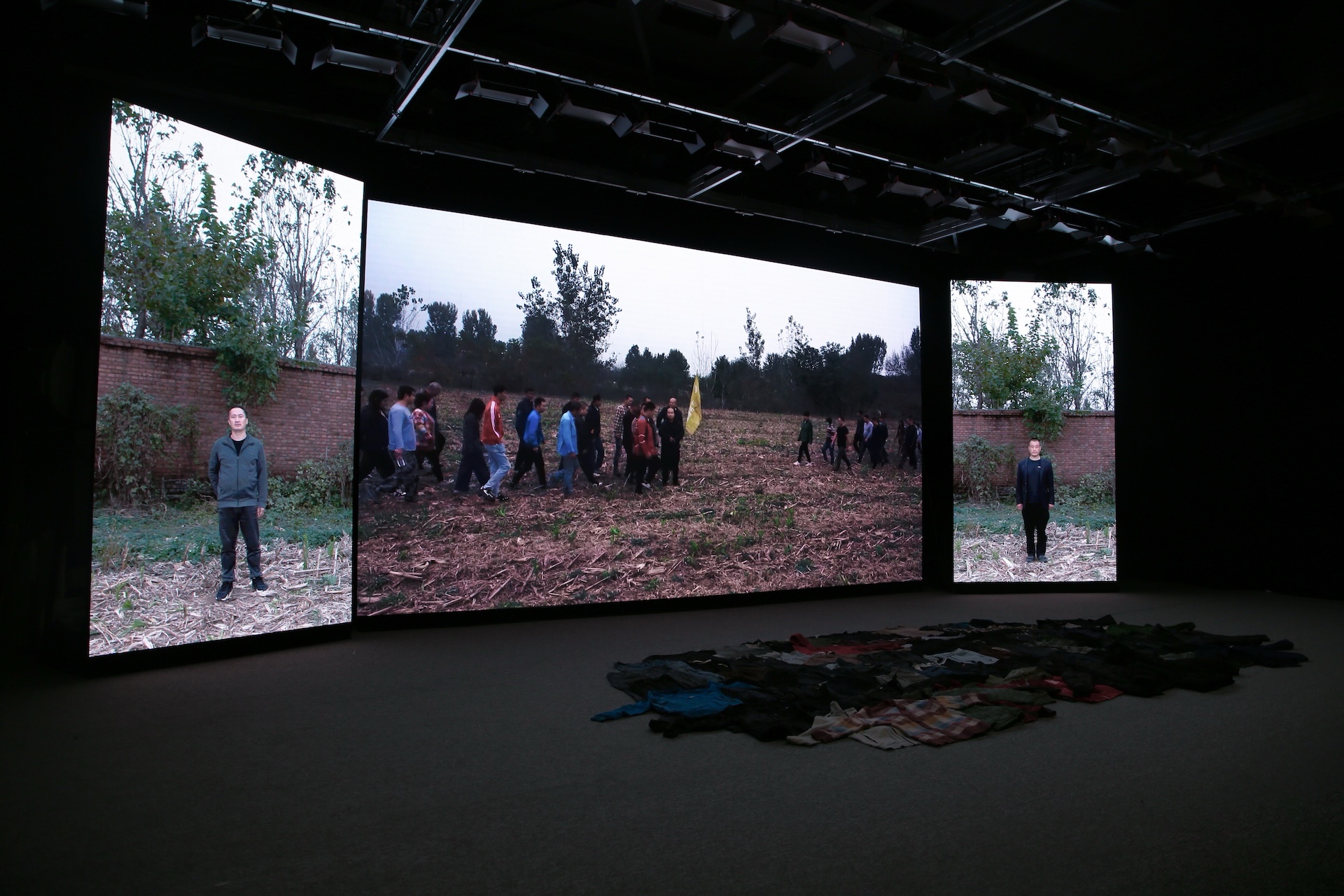 In a dark exhibition hall, three large screens show a group of people walking through an outdoor field, and a pile of clothes is laid out on the floor. From left to right, the left screen shows a man standing outdoors in front of a brick wall; the middle s