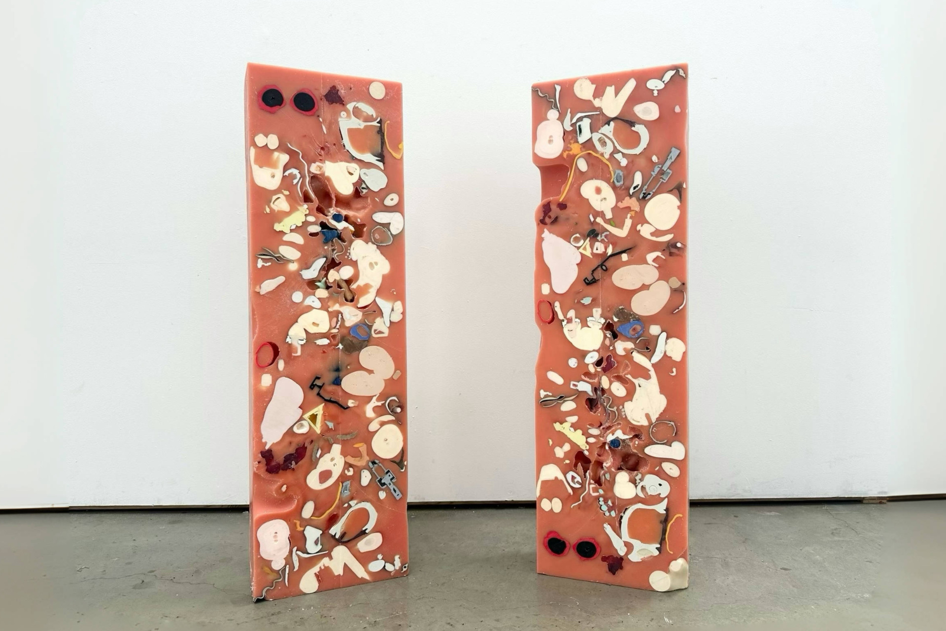 Two tall, narrow pink wax panels standing upright. Each panel contains embedded doll fragments being cross-sectioned in white, black, blue, red, and metallic tones. The surfaces feature irregular cutouts, bubbles, and textured details, giving it a strange 