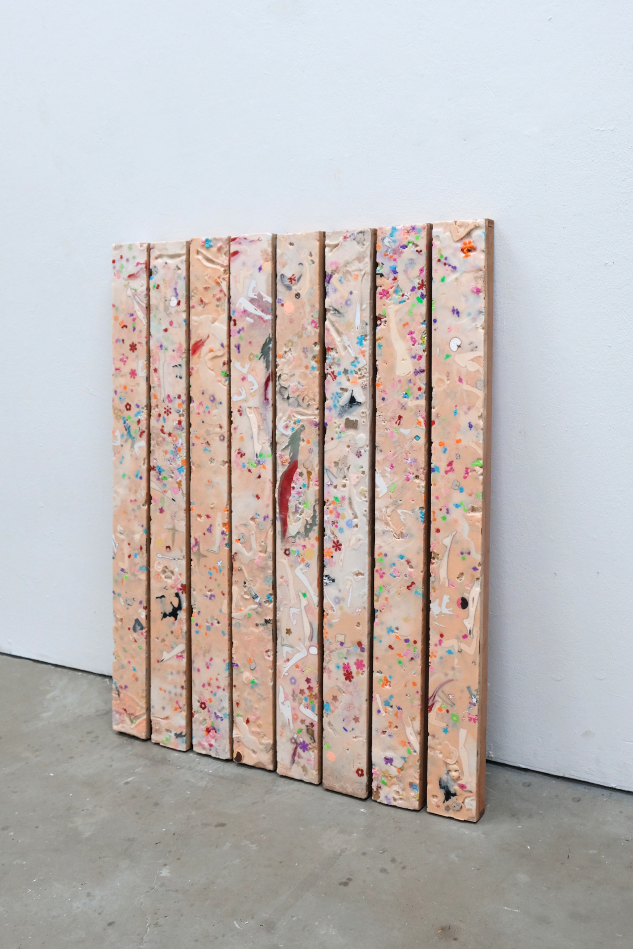 Eight narrow rectangular panels lean against a white gallery wall. The pale peach surfaces are embedded with colorful flecks, glitter, and small fragments of plastic dolls, creating a confetti-like pattern across the work. Arranged side by side, the panels