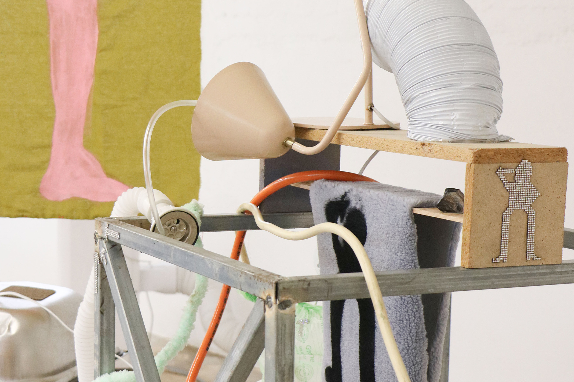 Detail of an installation featuring metal framing, cords, plush fabric, and found objects, including a lamp and a hose