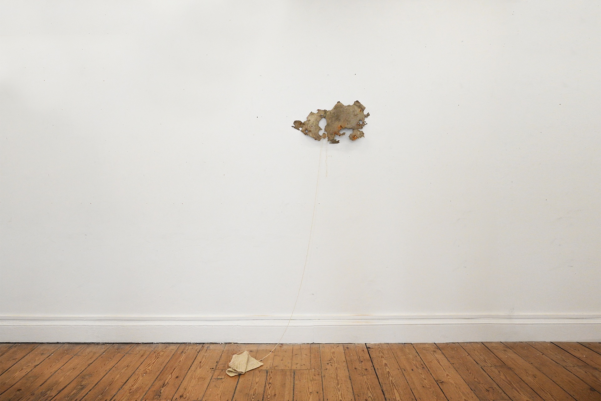 A rusted piece of scrap metal sits nailed to the wall. An unraveled piece of yarn hangs from its edges, and trails down to a knitted cream square lying crumpled on the floor. 