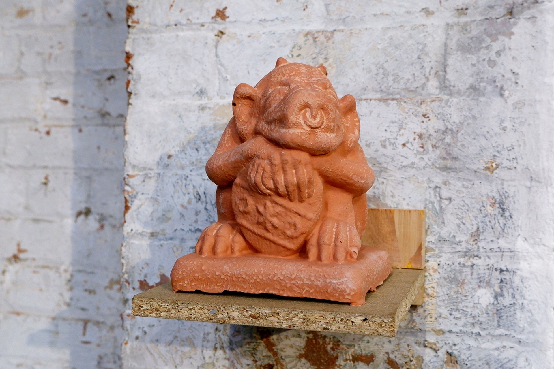 A grotesque of an ogre-like creature with a shield made from resin that looks like red brick, on a wooden shelf.