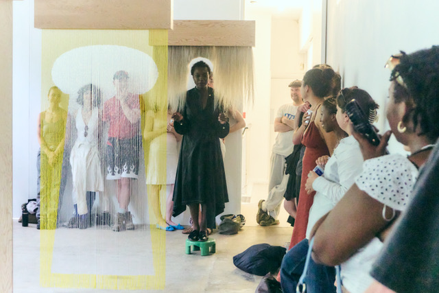 A photograph showing the artist Samra Mayanja doing a performance. She is standing on a small green stool in the middle of a brightly lit room dressed in a black, full-length dress. People stand alongside the walls, watching her.