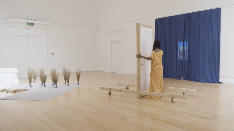 Installation view showing a woman dressed in yellow in front of a free-standing door. A blue curtain drapes one wall with a small projection in the middle, and dried grasses are displayed in bunches to the left.