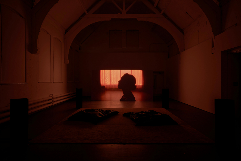 A dark room with a screen at the far end, showing the head of a person looking up, suffused with a warm orange colour.