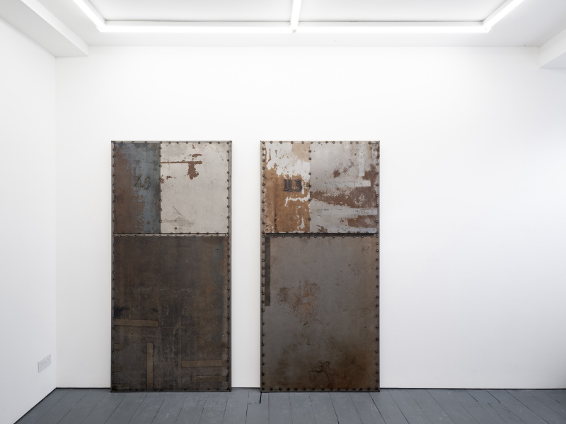 Installation view of an artwork by Ruskin tutor Racheal Crowther, showing two metal door shaped objects in a white room, dark grey with areas of rust.