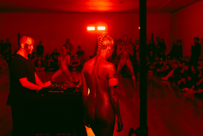 Image from a performance choreographed by artist Eddie Peake. Mostly nude performers bathed in red light express artistic notions of cultural and gender constructs.