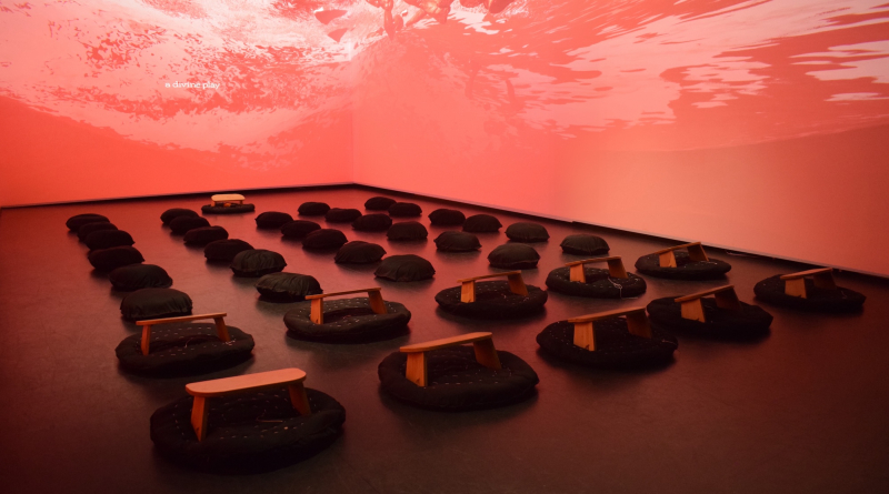 An image of the installation Your Rage is Sacred showing a dimly lit room with a swirling red pattern on the walls and cushions and small benches neatly arranged in the centre of the floor.