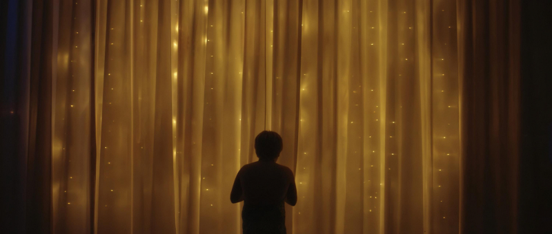 A still from a film by Eli Zuzovsky showing a child in front of a gold yellow curtain with light filtering through it.