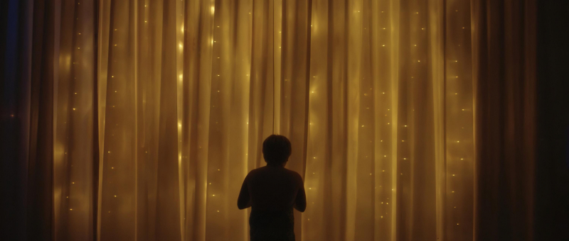 A still from a film by Eli Zuzovsky showing a child in front of a gold yellow curtain with light filtering through it.